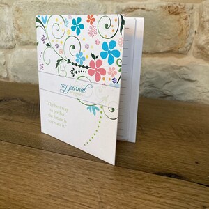 May include: A floral-patterned journal with the words "my journal" and a quote about creating the future. The cover features colorful flowers and green vines on a white background. The journal is open, revealing lined pages.