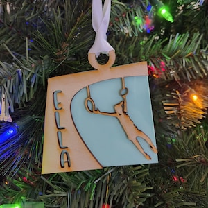 May include: A wooden Christmas ornament with a light blue background and a silhouette of a person on rings. The ornament is personalized with the name "ELLA" in brown lettering.