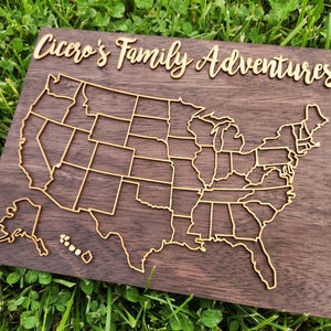 May include: A wooden map of the United States with a gold outline of the states. The map is mounted on a dark brown wooden board. The text "Cicero's Family Adventures" is in gold script above the map.