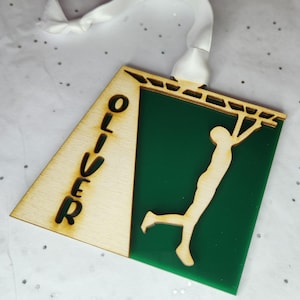 May include: A wooden ornament with a green background and a silhouette of a person hanging from a bar. The name "Oliver" is cut out of the wood on the left side of the ornament.