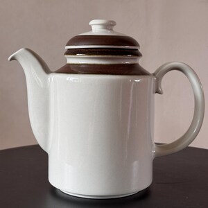 Op de afbeelding: Een keramische theepot met een gladde, gebroken witte behuizing, een gebogen handvat en een puntige tuit. Het deksel en de bovenrand van de theepot zijn versierd met een brede, donkerbruine band. Het deksel is voorzien van een kleine, witte knop.