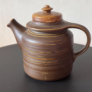 Op de afbeelding: Een donkerbruine keramische theepot met een cilindrisch lichaam, horizontale ribbeltexturen en een bijpassend deksel. De theepot heeft een gebogen tuit aan de linkerkant en een stevig handvat aan de rechterkant, met subtiele lichtbruine accenten.
