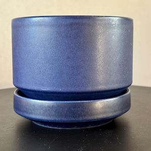 Op de afbeelding: Een cilindervormige, blauwe keramische bloempot met bijpassende schotel. De pot heeft een matte afwerking en een licht getextureerd oppervlak. De schotel is een ondiepe, cirkelvormige schaal die onder de pot past. Eenvoudig en modern ontwerp.