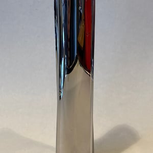 May include: A tall, polished silver vase with a sleek, modern design. The vase has a rectangular base that tapers upwards, with a rounded top. The reflective surface creates a play of light and shadow.