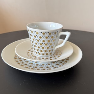 Puede incluir: Un juego de taza, platillo y plato blancos. La taza y los platos presentan un patrón repetido de círculos dorados y detalles azules. El juego se exhibe sobre una superficie oscura.