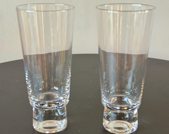 May include: Two clear, tall drinking glasses with a slightly flared top and a decorative base. The glasses are made of transparent glass and are set against a dark background. Ideal for water, juice, or cocktails.