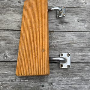 May include: A wooden handrail with a grooved surface and a light brown finish. It has two chrome-coloured metal brackets attached for mounting. The handrail is designed for use on stairs or other areas needing support.