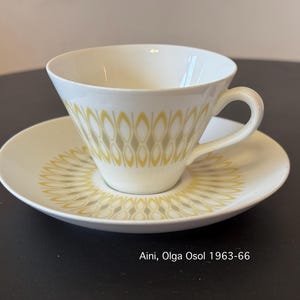 Puede incluir: Un juego de taza y platillo blancos con un diseño geométrico amarillo y gris. La taza tiene un asa y se apoya en un platillo a juego. El juego está etiquetado como "Aini, Olga Osol 1963-66".