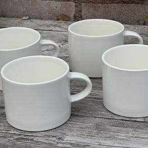 May include: Four white ceramic mugs with rounded bodies and attached handles are arranged on a weathered wooden surface. The mugs have a simple, minimalist design, suitable for coffee or tea.
