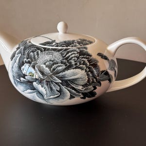 Könnte beinhalten: Eine weiße Teekanne aus Keramik mit einer großen, detaillierten schwarz-weißen Blumenillustration einer Pfingstrose. Die Teekanne hat einen abgerundeten Körper, einen geschwungenen Ausguss, einen passenden Deckel mit Knauf und eine glatte Oberfläche.