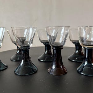 May include: Set of six clear glass cordial glasses with dark gray stems and bases. The glasses have a flared bowl shape and sit on a short, rounded stem. The glasses are arranged on a dark surface.