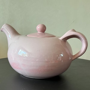 Puede incluir: Una tetera de cerámica rosa con un cuerpo redondeado y un diseño sencillo. La tetera tiene una tapa con un pomo en la parte superior y un asa en el lateral.