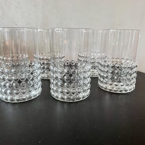 May include: Set of six clear glass tumblers. The bottom of each glass features a textured, bubble-like design. The glasses are cylindrical with a smooth, clear upper portion. The glasses are arranged on a dark surface.