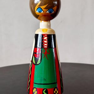 May include: A vintage wooden doll with a round head and a painted face featuring blue eyes and brown hair. The doll wears a dress with a green skirt, red accents, and black bodice. The doll is approximately 7 inches tall.