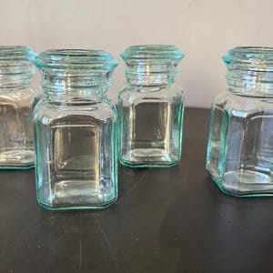 Peut inclure: Quatre bocaux en verre transparent avec une légère teinte turquoise. Les bocaux ont une base carrée et un dessus arrondi et strié. Les bocaux sont vides et reposent sur une surface sombre.