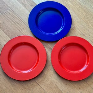 Puede incluir: Tres platos coloridos: uno azul y dos rojos. Los platos son redondos con un borde ligeramente elevado. Los platos están sobre una superficie de madera de color marrón claro.