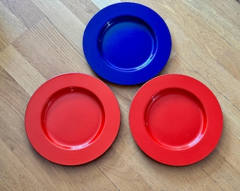 May include: Three colourful plates: one blue and two red. The plates are round with a slightly raised rim. The plates are on a light brown wooden surface.