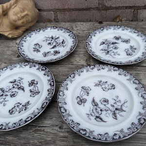 Puede incluir: Cuatro platos de cerámica blanca con un patrón floral oscuro. Los platos tienen un borde festoneado y un borde detallado. El diseño floral incluye flores y hojas. Los platos están dispuestos sobre una superficie de madera.