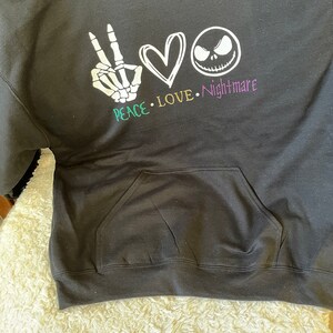 K&ouml;nnte beinhalten: Schwarzer Hoodie mit einem wei&szlig;en Peace-Zeichen, einem Herzen und einem Jack-Skellington-Gesicht. Der Text "Peace. Love. Nightmare" ist unter dem Grafikdruck.