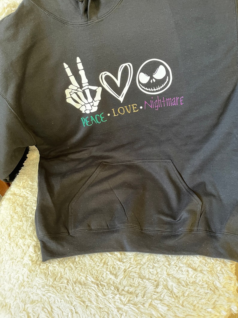 K&ouml;nnte beinhalten: Schwarzer Hoodie mit einem wei&szlig;en Peace-Zeichen, einem Herzen und einem Jack-Skellington-Gesicht. Der Text "Peace. Love. Nightmare" ist unter dem Grafikdruck.