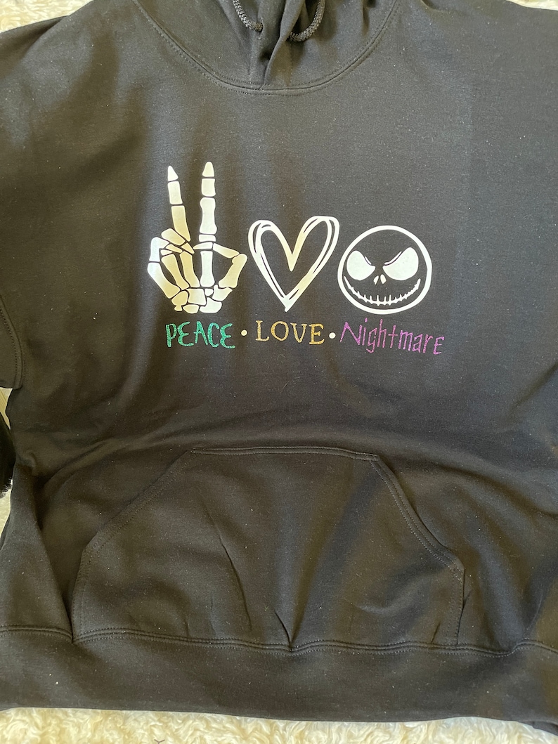 K&ouml;nnte beinhalten: Schwarzer Hoodie mit einem wei&szlig;en Peace-Zeichen, einem Herzen und einer Grafik von Jack Skellington. Der Text "Peace Love Nightmare" ist unter der Grafik gedruckt.