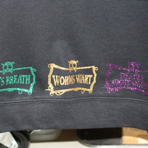 K&ouml;nnte beinhalten: Schwarzer Pullover mit drei glitzernden B&uuml;gelbildern. Die Patches haben die Form eines Banners mit einem Totenkopf und gekreuzten Knochen oben. Der Text auf den Patches lautet "Frog's Breath", "Worms Wart" und "Dragon's Rot Gut".