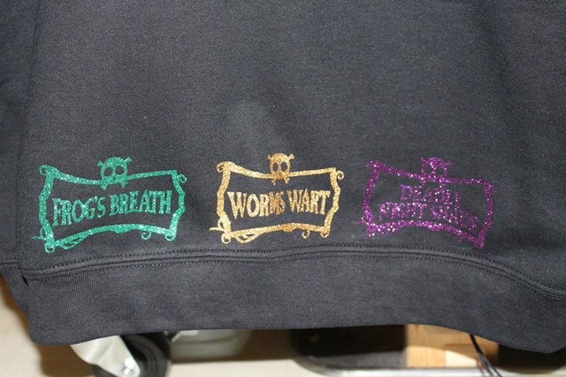 K&ouml;nnte beinhalten: Schwarzer Pullover mit drei glitzernden B&uuml;gelbildern. Die Patches haben die Form eines Banners mit einem Totenkopf und gekreuzten Knochen oben. Der Text auf den Patches lautet "Frog's Breath", "Worms Wart" und "Dragon's Rot Gut".