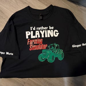 May include: Black t-shirt with white text that reads "I'd rather be playing Farming Simulator". The shirt also has a green and black tractor graphic and the words "Ginger Nuts" on the sleeves.