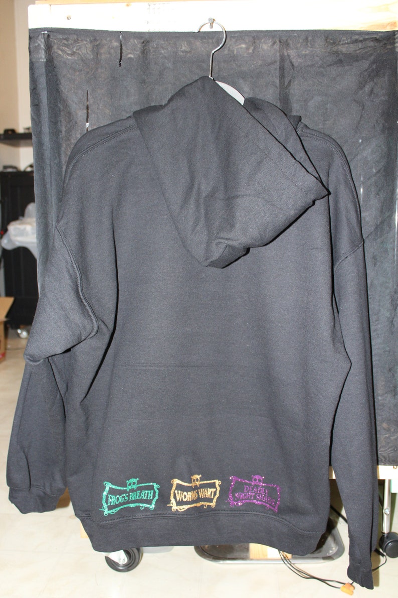 K&ouml;nnte beinhalten: Schwarzer Kapuzenpullover mit drei gestickten Patches am unteren Rand. Die Patches sind in Gr&uuml;n, Gold und Lila und zeigen den Text "Frog's Breath", "Work's Want" und "Death Don't Stop".