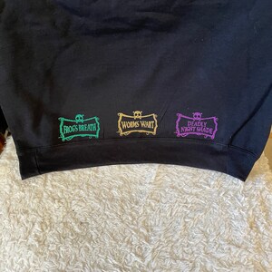 K&ouml;nnte beinhalten: Schwarzer Pullover mit drei gestickten Patches. Die Patches sind rechteckig und haben ein Totenkopf- und Knochenkreuz-Design. Der Text auf den Patches lautet "Frog's Breath", "Worm's Wart" und "Deadly Nightshade".