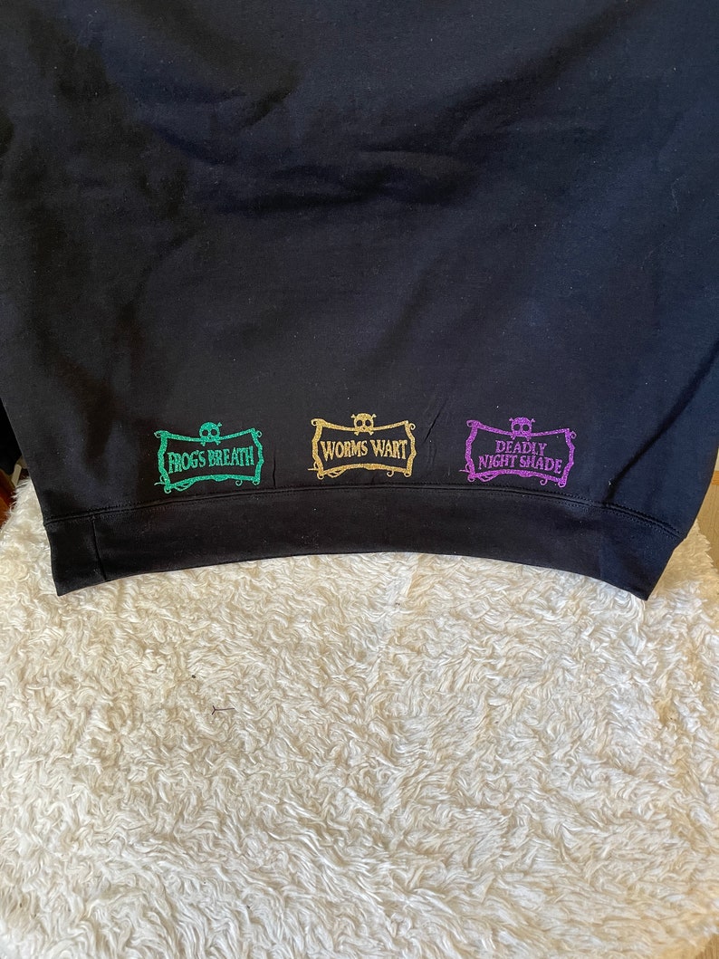 K&ouml;nnte beinhalten: Schwarzer Pullover mit drei gestickten Patches. Die Patches sind rechteckig und haben ein Totenkopf- und Knochenkreuz-Design. Der Text auf den Patches lautet "Frog's Breath", "Worm's Wart" und "Deadly Nightshade".