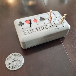 May include: A grey 3D printed Euchre game board with black and red card suit symbols and a black peg. The board has a grey lid with the word "EUCHRE" printed on it. A silver metal token with the word "EUCHRE" and card suit symbols is in the foreground.