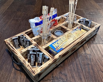 May include: A wooden tray with compartments for holding s'mores ingredients. The tray has a burnt wood finish and includes chocolate bars, graham crackers, marshmallows, skewers, and a jar of honey.