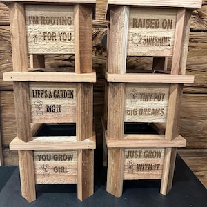 Könnte beinhalten: Vier gestapelte Holzpflanzgefäße mit graviertem Text und Illustrationen. Der Text enthält Sätze wie „I'm rooting for you“, „Raised on sunshine“ und „Tiny pot, big dreams“. Die Pflanzgefäße sind aus hellbraunem Holz.