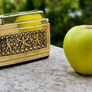Puede incluir: Un encendedor dorado y adornado con un diseño detallado y una manzana verde sobre una superficie gris claro.