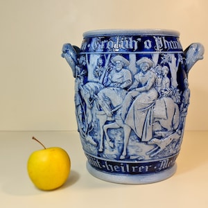May include: Blue and white ceramic pitcher with a relief design of people riding horses and a dog. The pitcher has a handle on each side and the text "Grädlüh' o Phom" on the front.