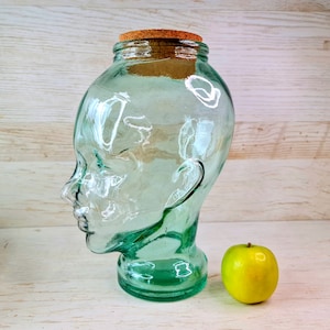 以下が含まれることがあります： コルクの蓋が付いた緑色のガラス製の頭の形をした瓶。瓶は白い木製の表面に置かれ、緑色のリンゴの隣にあります。