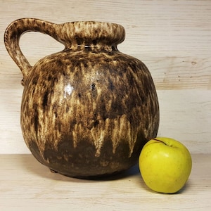 Puede incluir: Un jarrón de cerámica marrón y crema con una boca ancha y un asa. El jarrón tiene un esmalte moteado y está sentado sobre una superficie de madera junto a una manzana verde.