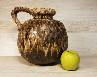 May include: A brown and cream ceramic vase with a wide mouth and a handle. The vase has a mottled glaze and is sitting on a wooden surface next to a green apple.