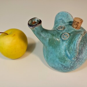 May include: A teal blue ceramic teapot shaped like an elephant with a cork stopper. The teapot has a textured surface and a small spout.