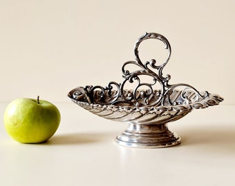 May include: A silver serving dish with a decorative handle and a swirl design. The dish is on a pedestal and is oval shaped.