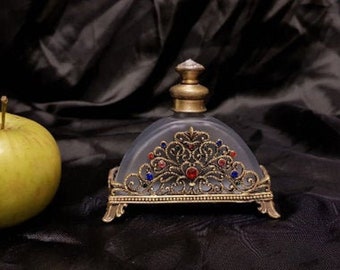 May include: A frosted glass bottle with a gold metal top and ornate design. The bottle is on a gold metal stand with a decorative design. The bottle has red and blue jewels.
