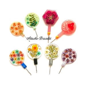 May include: Assortment of colorful round and hexagonal resin charms with pressed flowers and leaves. Each charm has a silver pin attached to the back. The charms are arranged on a white surface. "Attache-Bracelet" is written in black on the surface.