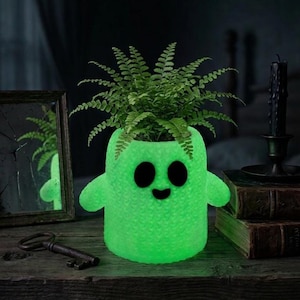 May include: A glowing neon green planter shaped like a ghost with a textured, knit-like surface. The ghost has two oval black eyes and a small black mouth. A vibrant green fern plant is growing out of the top opening of the ghost-shaped container.