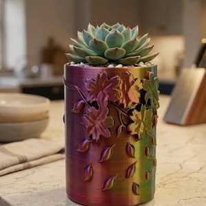 May include: A cylindrical planter with a rainbow gradient finish featuring embossed leaf and floral patterns on the exterior. A green succulent plant is potted inside, surrounded by small white pebbles at the top of the container.