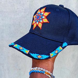 Könnte beinhalten: Eine schwarze Baseballkappe mit einem bunten Perlensonnen-Design auf der Vorderseite. Der Schirm der Kappe ist ebenfalls mit Perlen verziert.