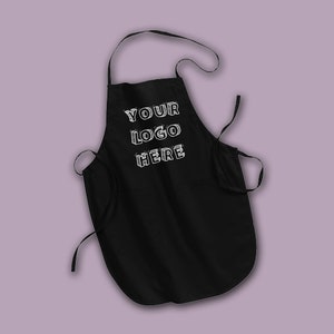 Puede incluir: Delantal negro con una correa de cuello ajustable y dos lazos largos. El delantal tiene un bolsillo grande en la parte delantera y texto blanco que dice "YOUR LOGO HERE".