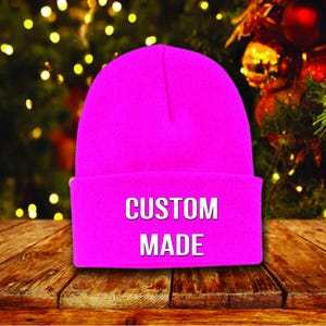 May include: A bright pink beanie with the words "CUSTOM MADE" in white block letters. The hat is folded at the brim and sits on a wooden surface. The background features a blurred Christmas tree with lights and ornaments.