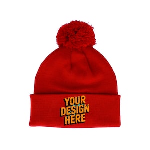 Op de afbeelding: Een felrode beanie met een bijpassende pompon. De omgeslagen rand toont de tekst "YOUR DESIGN HERE" in een opvallend, gestileerd lettertype. Dit winteraccessoire is aanpasbaar.
