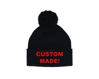 Può includere: Un berretto nero con un pompon nero abbinato. Il risvolto mostra il testo rosso "CUSTOM MADE!". Questo cappello invernale è un accessorio versatile per il clima freddo.